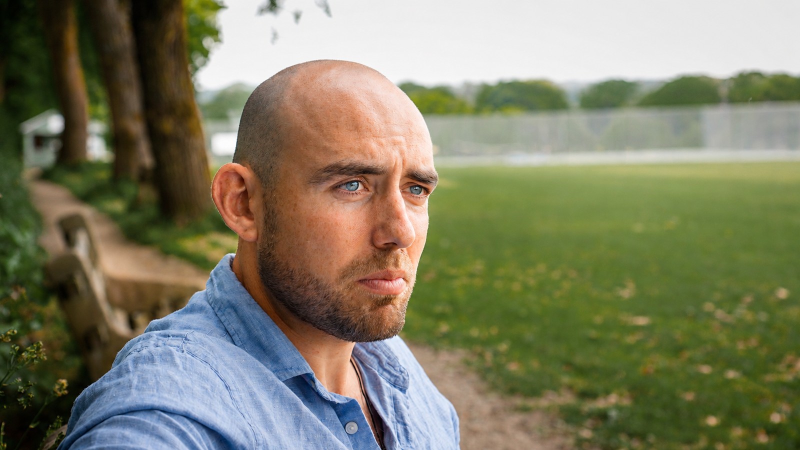 Jack looks off to the side, sitting on a bench next to a green field.