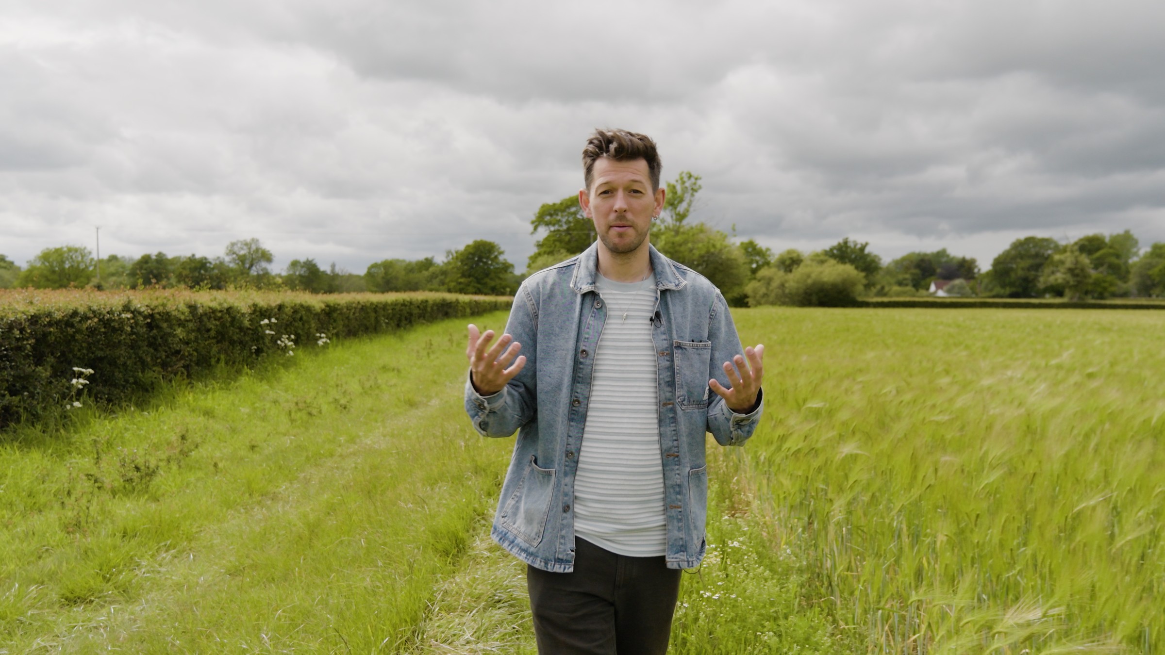 Josh Westover stands in a green field in a denim jacket and talks to the camera.