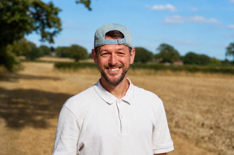 Josh Westover smiles at the camera, wearing a cap, in a sunlit field.